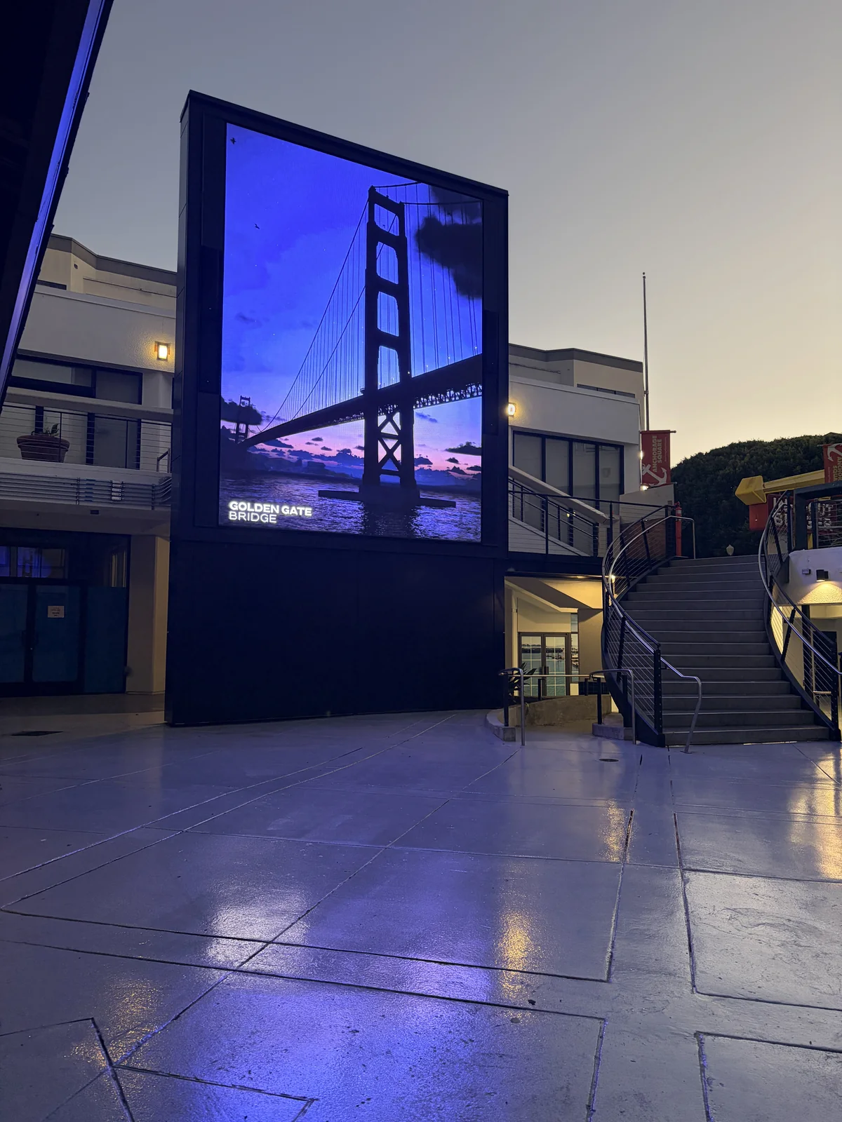 The tall portrait LED canvas in the Anchorage Square plaza showing the Golden Gate Bridge at sunset, its light reflecting off the wet paving