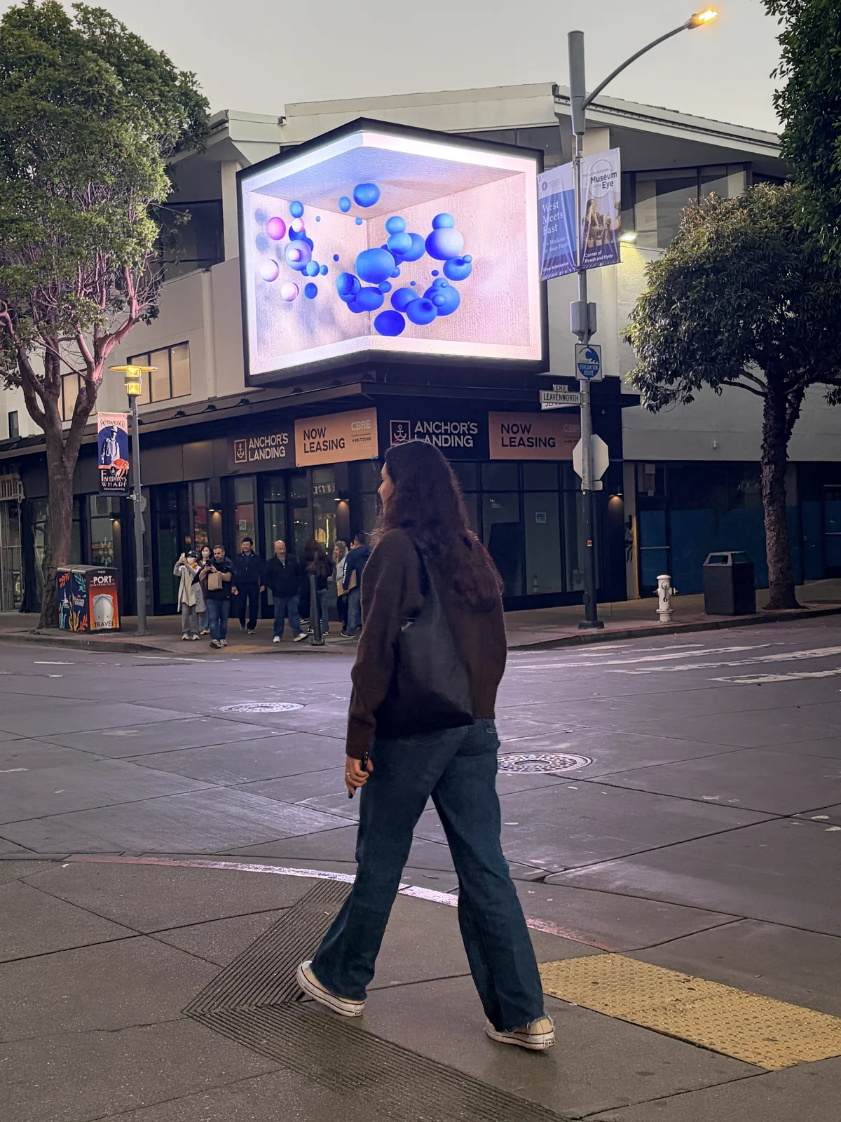The corner LED volume at Anchorage Square running an anamorphic scene of blue spheres suspended inside an open box, seen from the sidewalk as a passer-by walks past
